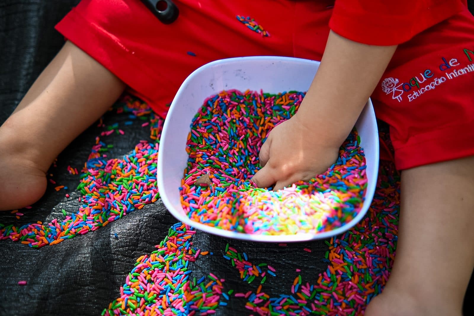 Mãos de uma criança e de uma educadora mexendo em uma bacia de arroz colorido