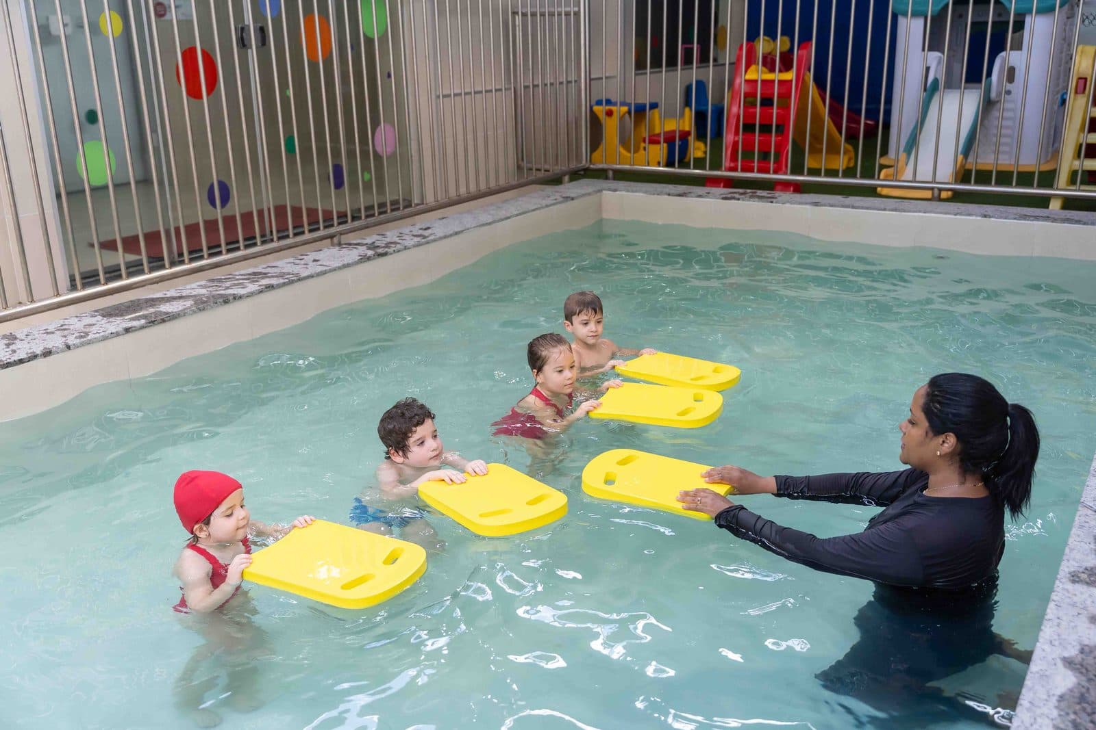 Professora na piscina com quatro crianças apoiadas em pranchas amarelas