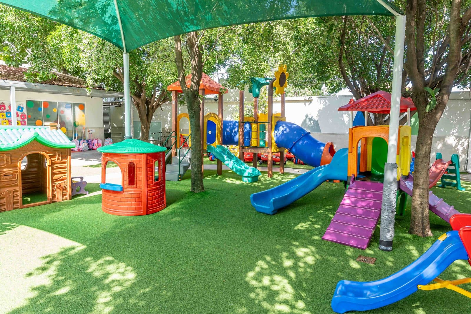 Playground coberto por árvores, com casinhas, escorregadores e piso emborrachado verde