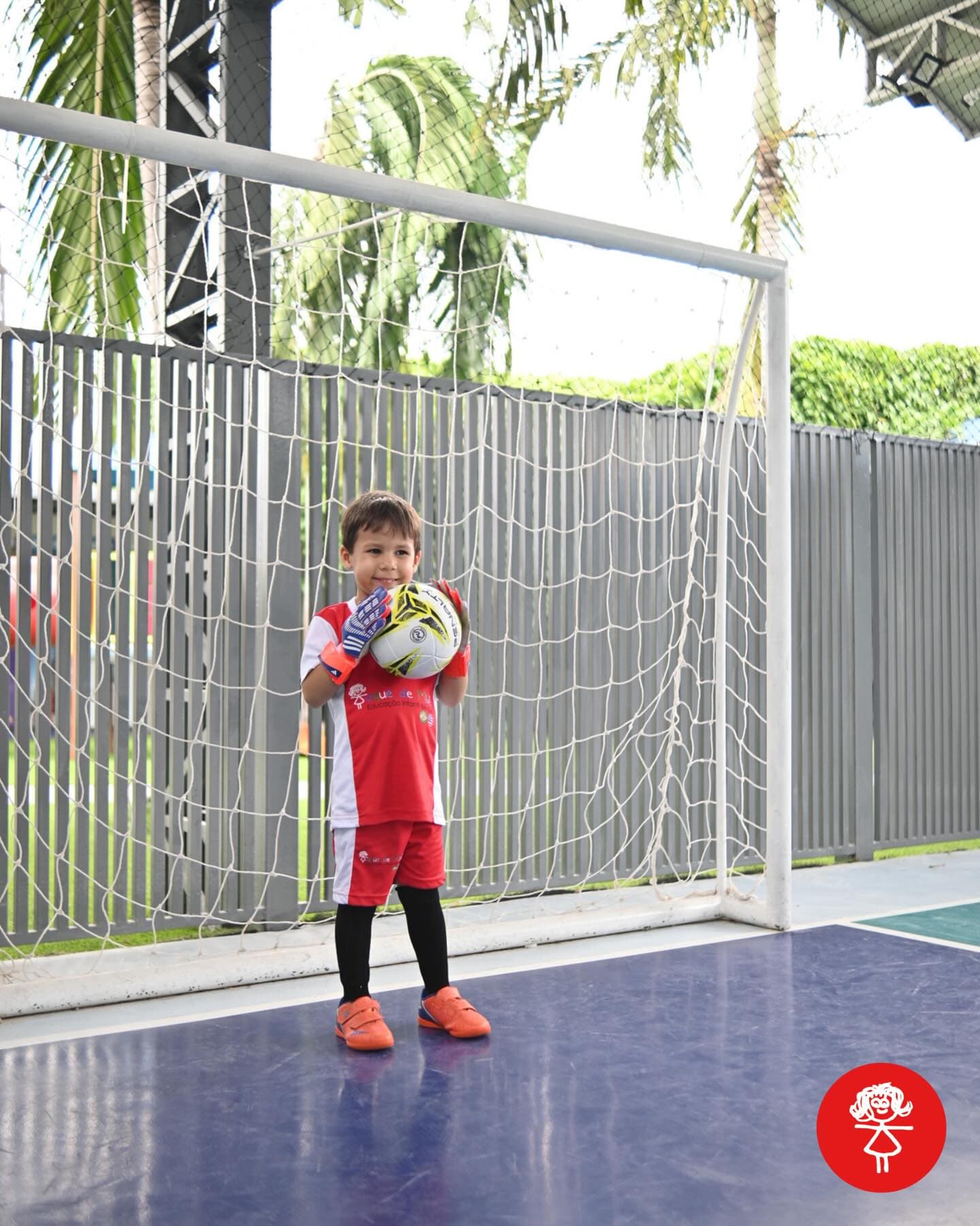 Criança de uniforme vermelho em frente ao gol, segurando a bola de futsal