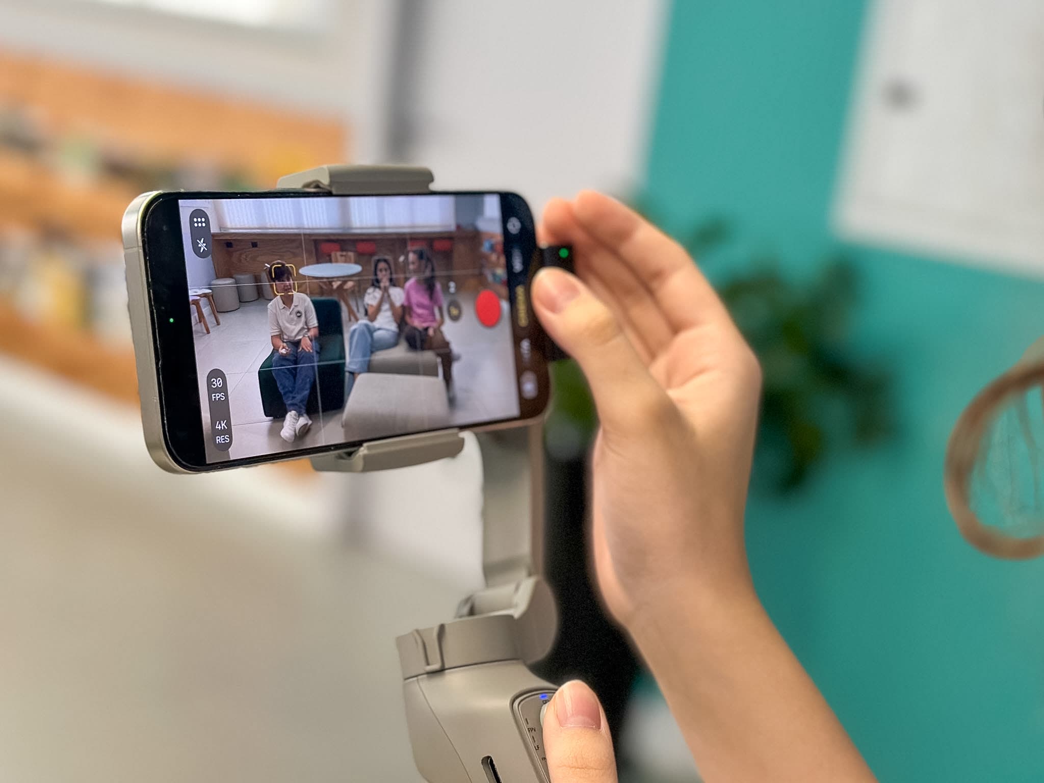 Mãos de uma aluna segurando o celular num estabilizador, gravando três estudantes sentados num sofá da escola
