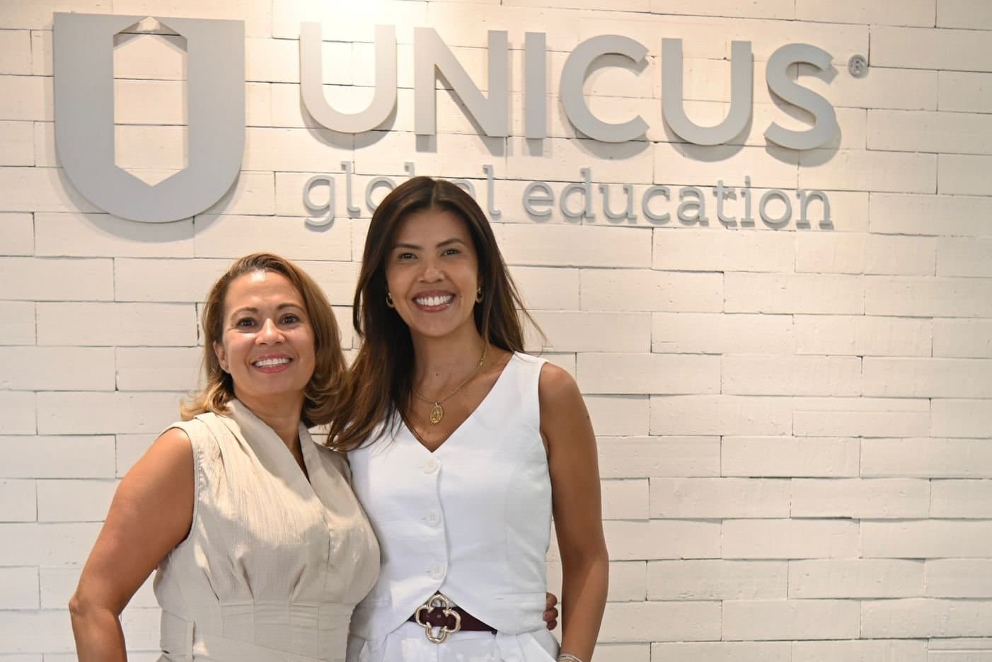 Duas educadoras lado a lado diante da parede com o logo do Colégio Unicus