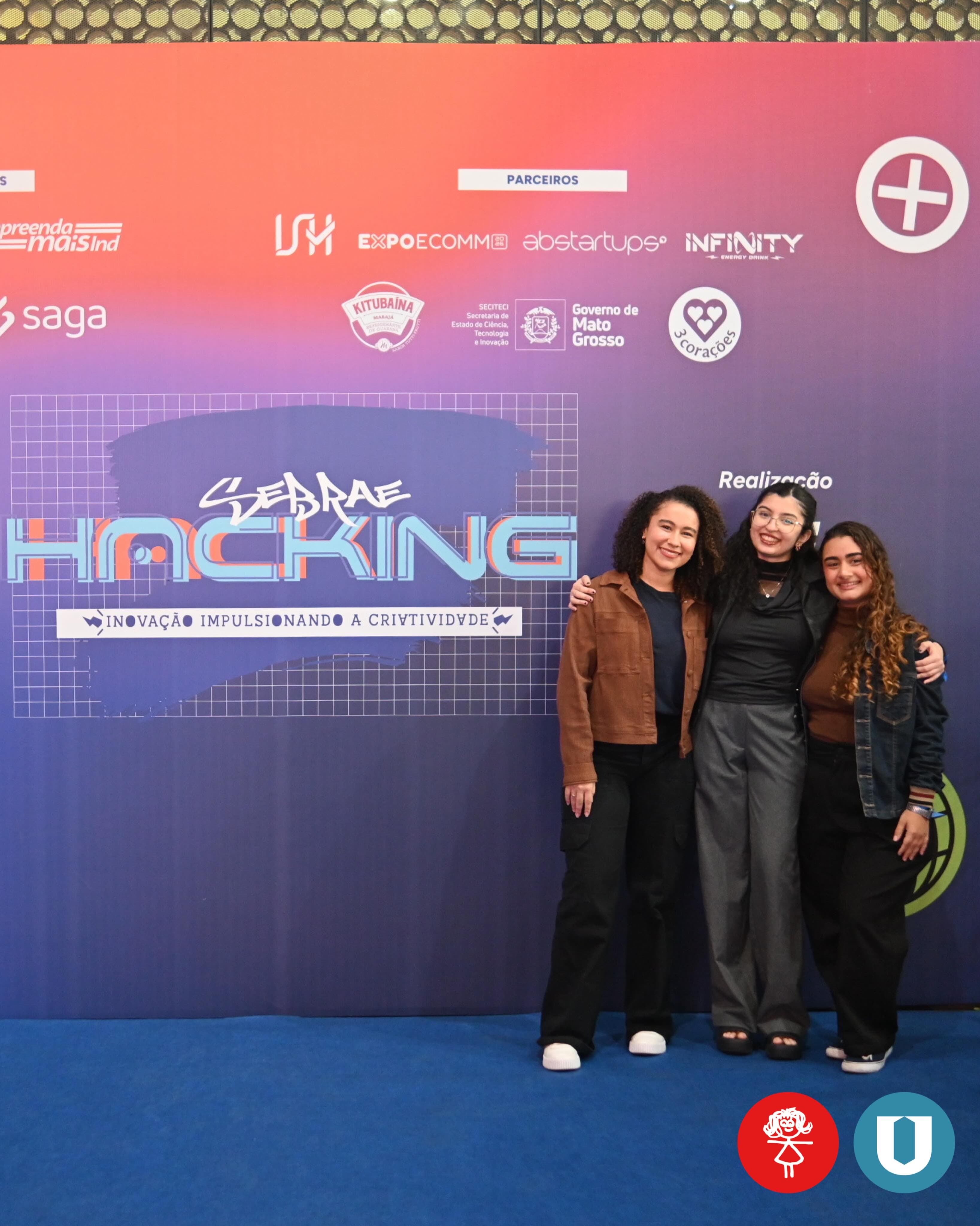 Três integrantes da equipe da escola diante do painel do Sebrae Hacking