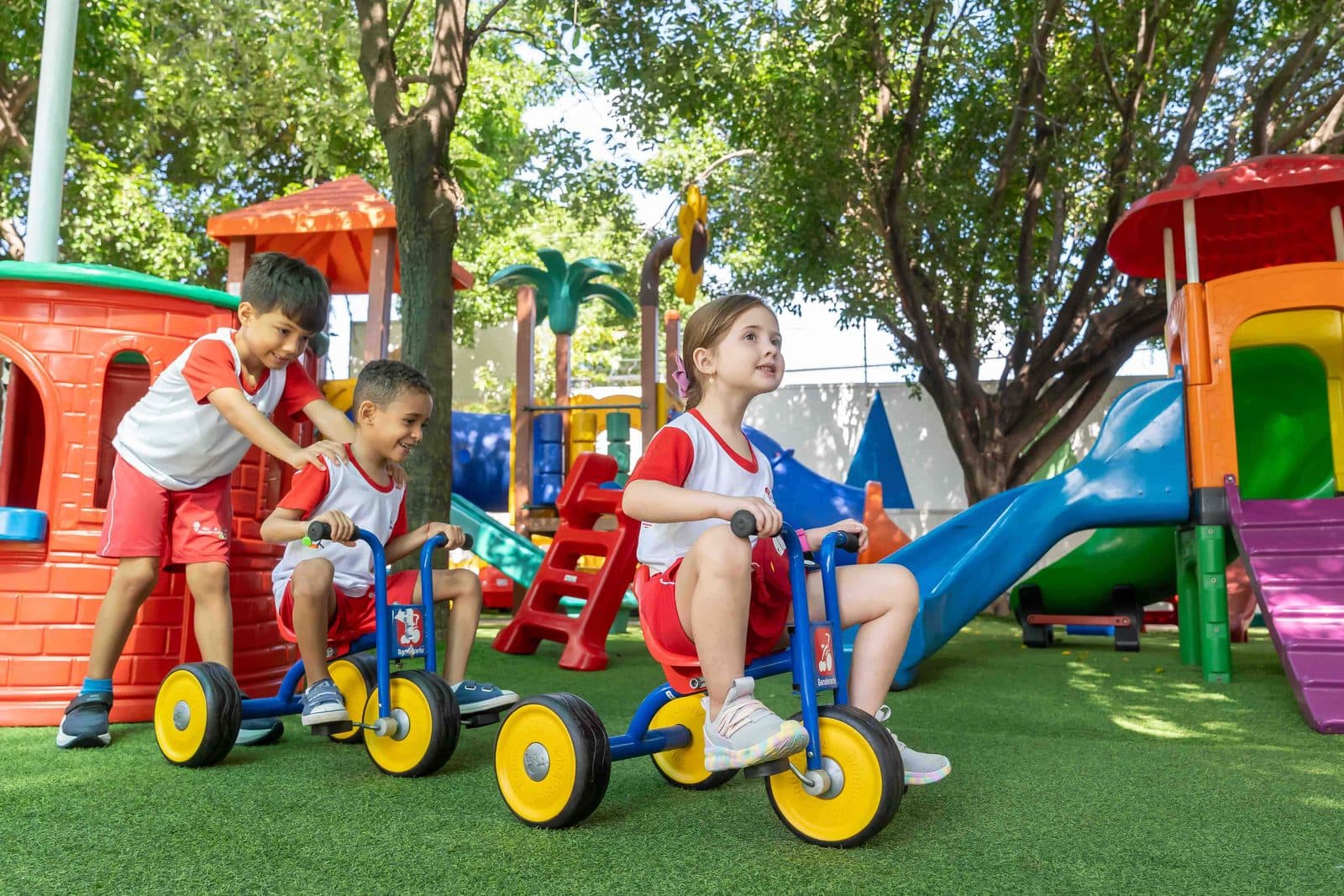 Crianças andando de triciclo no playground gramado, à sombra das árvores
