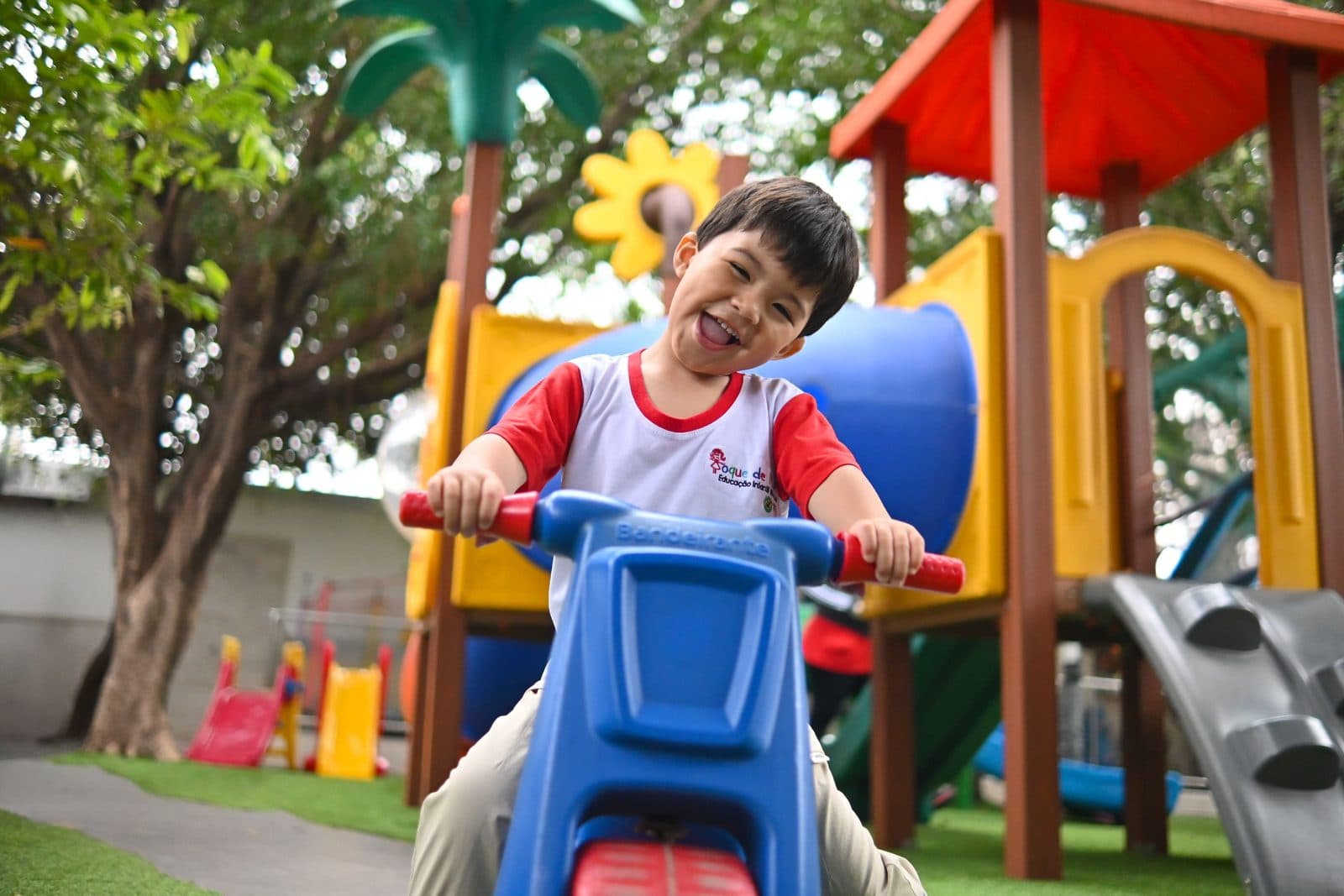 Criança sorrindo em cima do triciclo, no playground colorido da escola