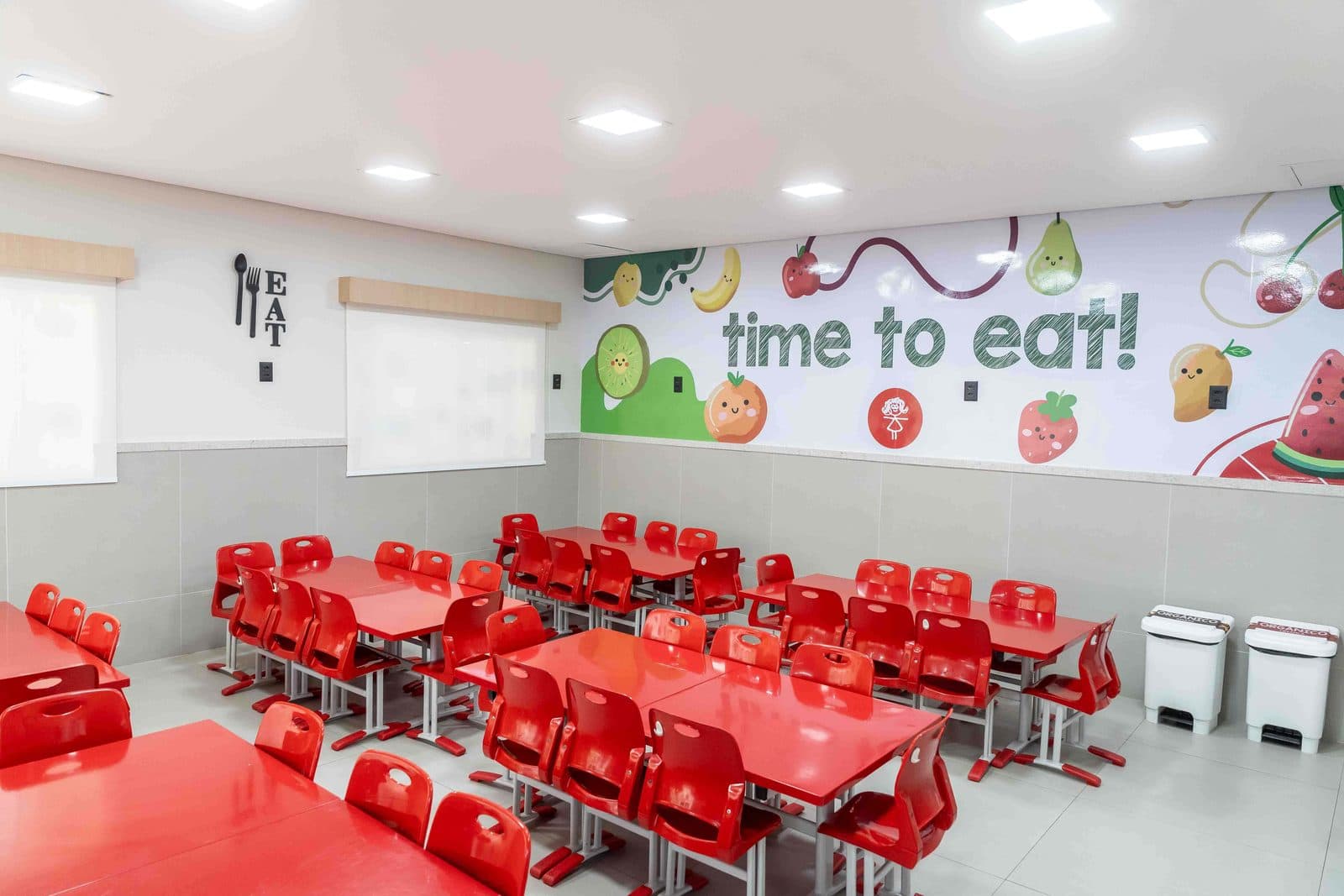 Refeitório da escola, com mesas e cadeiras vermelhas e mural ilustrado de frutas